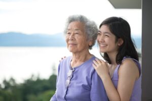 Elderly Asian woman and granddaughter smiling with mountains and water in the background.